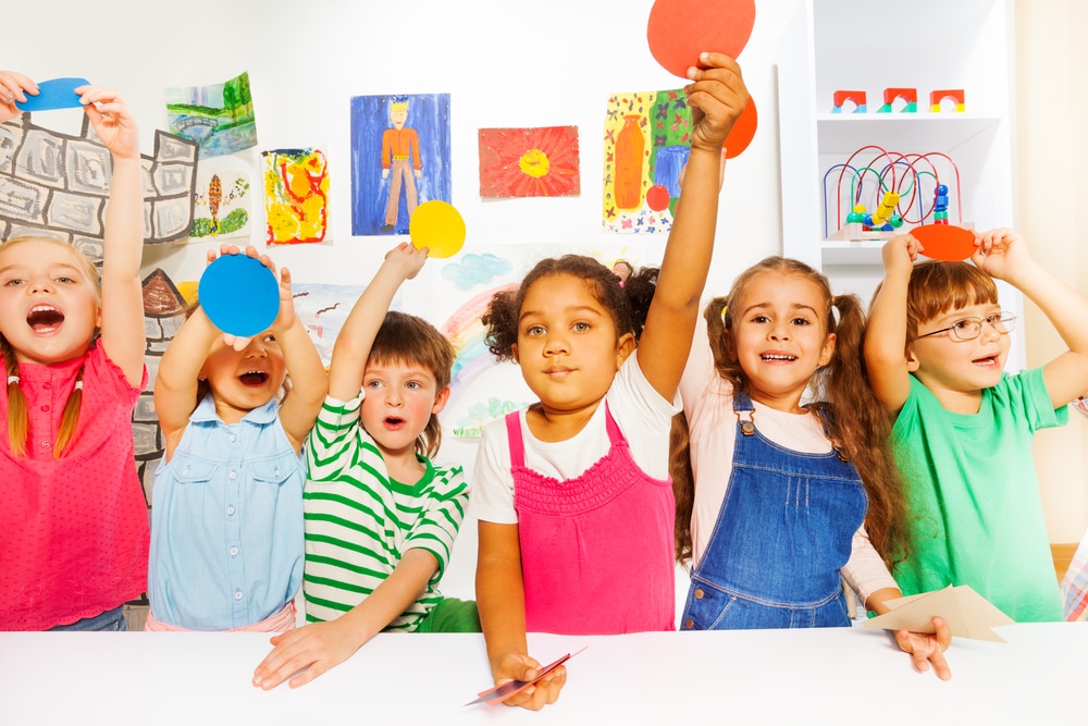 Group of happy boys and girls in kindergarten holding color cardboard shapes and looking at camera